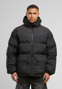 Giacca a vento nera con collo alto, chiusura con zip, due tasche frontali e polsini a costine. Il design presenta texture di pannelli a contrasto.