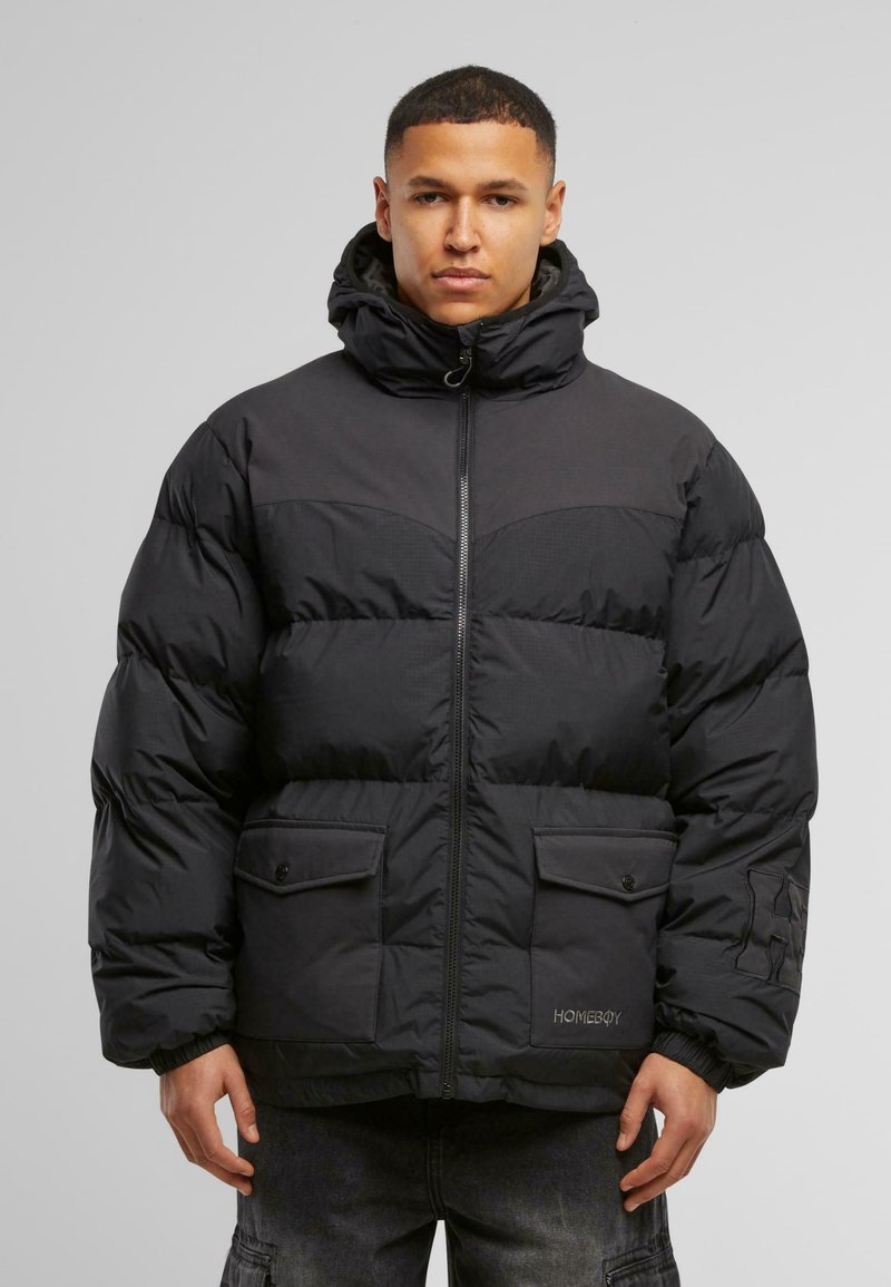 Giacca a vento nera con collo alto, chiusura con zip, due tasche frontali e polsini a costine. Il design presenta texture di pannelli a contrasto.