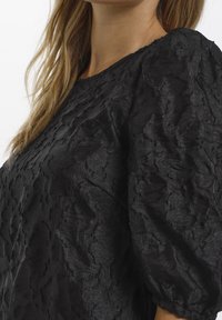 Kvinna iklädd en svart blus med texturerade puffärmar och rund halsringning, synlig från nacken till mitten av bålen.