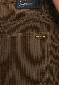 Pantalons en velours côtelé marron avec des côtes verticales, une étiquette en tissu noir avec un design en relief, et une petite étiquette "Volcom" sur la poche.