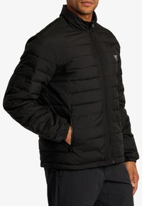 Sort pufferjakke med høj krave, horisontal quiltning, lynlås foran og sidelommer. Har elastiske manchetter og et logo på brystet.