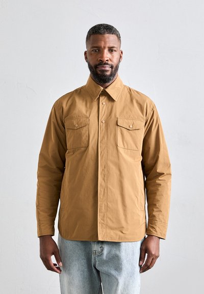 Veste marron à boutons avec deux poches poitrine, manches longues et col. Tissu lisse, coupe décontractée, associée à un jean bleu clair.