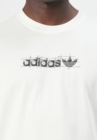 White cotton t-shirt featuring a black printed logo with a retro design, including the name "adidas" and the classic trefoil emblem.