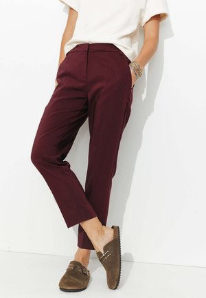 Pantalons taillés bordeaux à jambe droite, avec poches latérales et fermeture éclair. Associés à des chaussures slip-on en suède marron avec des accents cloutés.