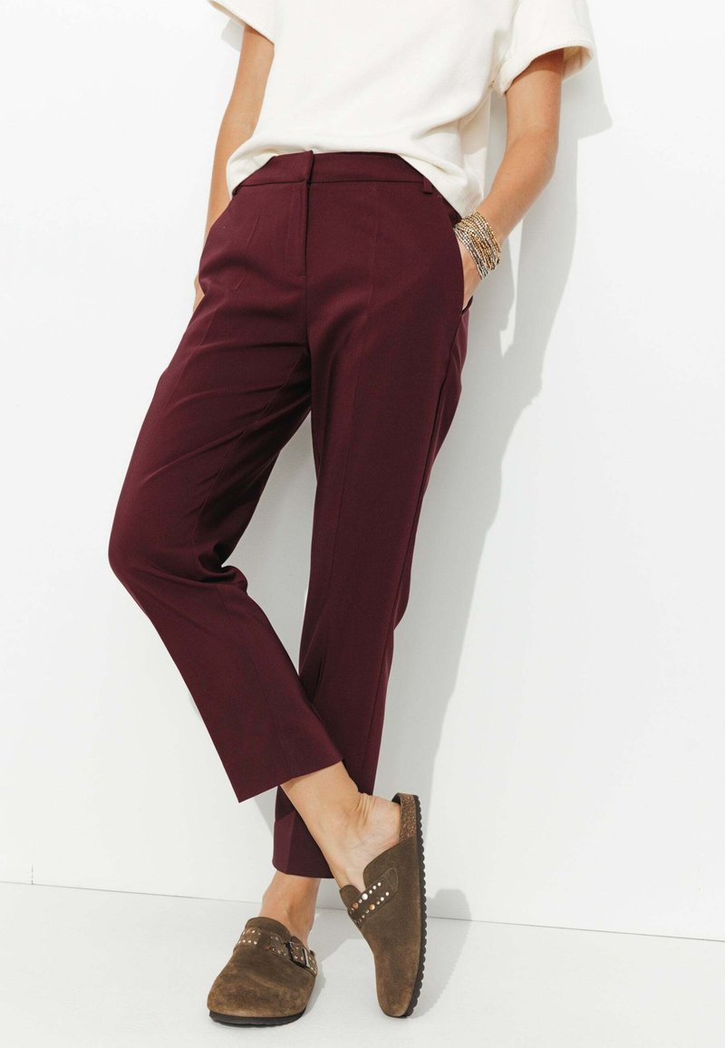 Pantalons taillés bordeaux à jambe droite, avec poches latérales et fermeture éclair. Associés à des chaussures slip-on en suède marron avec des accents cloutés.