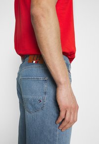 Jeans de mezclilla azul claro con un corte ajustado y costuras en contraste. Presenta un parche de cuero marrón con el logotipo y un pequeño detalle de bandera en el bolsillo.