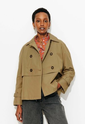 Femme portant un trench-coat beige croisé sur un chemisier rouge à fleurs et un jean gris foncé, debout avec une main dans la poche.