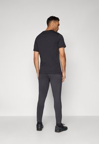 Svart bomull T-skjorte og mørkegrå tights, tettsittende design, glatt tekstur. Modell iført svarte treningssko. Bakfra, ensfarget bakgrunn.