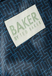 Modrý vzorovaný textil s bílou visačkou se zeleným nápisem "BAKER by TED BAKER." Design zahrnuje geometrické tvary a linie.