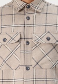 Beige flanellen shirt met een geruit patroon in zwart en crème, met twee borstzakken en donkere knopen. De textuur lijkt zacht te zijn.