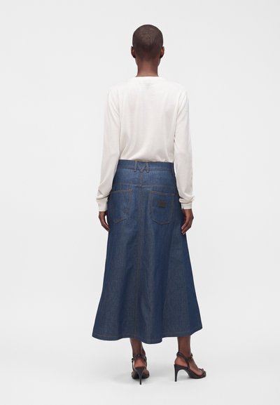 Femme vue de dos portant une chemise blanche à manches longues, une jupe midi en denim bleu foncé et des sandales noires à talons hauts.