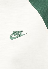 Λευκό ύφασμα με κέντημα πράσινου λογότυπου Nike και πράσινο ύφασμα στην πάνω δεξιά γωνία.