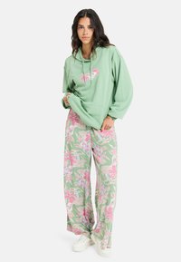 Felpa verde con design floreale rosa, vestibilità morbida e tasca frontale; abbinata a pantaloni floreali a gamba larga con colori simili e scarpe bianche.