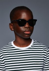 Čierne slnečné okuliare Ray-Ban so štvorcovými rámami, nosené s bielym a zeleným pruhovaným tričkom s malým logom "BOSS" na hrudi.