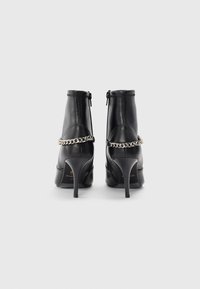 Bottes noires en cuir à cheville avec une finition lisse, talons aiguilles hauts et un accent en chaîne argentée autour de la cheville. Fermeture éclair à l'arrière.