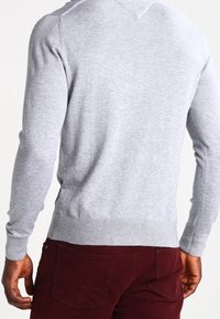 Grauer Pullover mit langen Ärmeln, gerippten Bündchen und Saum, getragen über burgunderfarbenen Hosen. Verfügt über ein einfaches, strukturiertes Strickdesign.