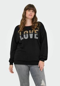 Sieviete ar viļņainiem brūniem matiem, ģērbta melnā džemperī ar uzrakstu "LOVE" leopardu printā un pelēkos burtiem, apvienojot ar gaiši pelēkiem plēstiem džinsiem.