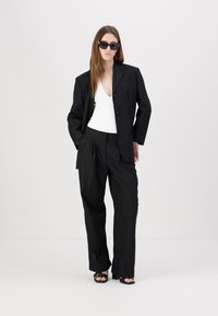 Svart oversized blazer med skjæring ved kragen, kombinert med høytlivede svarte bukser og en hvit bodysuit. Tilbehøret inkluderer svarte solbriller.