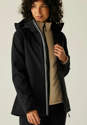 Kobieta z kręconymi włosami, ubrana w czarną wodoodporną kurtkę na beżowym polarze, zapinająca zamek kurtki, stojąca na jednolitym tle.