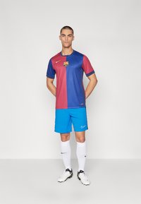 Maglia del FC Barcelona con metà rossa e metà blu, maniche corte e logo. Indossata con pantaloni corti blu brillanti, calzettoni bianchi e scarpini da calcio neri.