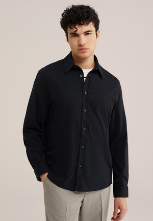 Giovane uomo con capelli ricci che indossa una camicia nera con bottoni sopra una maglietta bianca, mani parzialmente nelle tasche, in piedi davanti a uno sfondo beige.