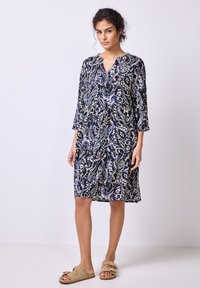 Laivansinis mekko, jossa on paisley-kuvio, polvipituinen, V-mallinen kaula-aukko, 3/4 hihat, kevyttä kangasta, yhdistetty vaaleisiin sandaaleihin.
