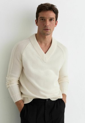 Muž v krémovom svetri s výstrihom do V a čiernych nohaviciach, stojaci s rukami v vreckách pri bielej stene.