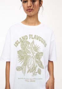 Weißes Baumwoll-T-Shirt mit einem grünen botanischen Grafik- und Textdesign. Es enthält Elemente wie Kakaopods und tropische Blätter.