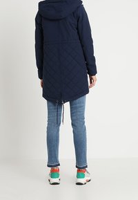 Granatowy parka z pikowanym dolnym sekcją, regulowanym paskiem w talii i kapturem, zestawiona z dopasowanymi niebieskimi dżinsami i kolorowymi sneakersami.