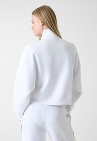 Weißes Sweatshirt mit hohem Kragen, abgesenkten Schultern und glatter Textur, angeschnitten. Kombiniert mit passenden weißen Hosen.