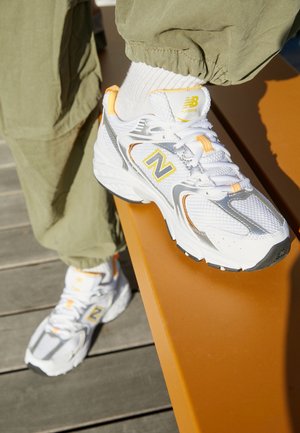 Hvide og sølvfarvede New Balance løbesko med gule accenter, båret med hvide sokker og olivengrønne bukser, fod hvilende på en orange bænk.