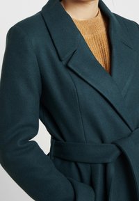 Mørk teal bælteuld frakke båret over en tekstureret kamel-farvet striktrøje, der viser overkroppen og armen.