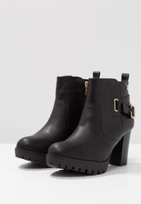 Et par sorte læder ankelstøvler med blokhæle, sidezipper, dekorativ rem og guldspænde mod en simpel lys baggrund.