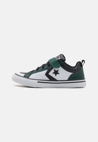 Zapatilla con parte superior de piel blanca, detalles en negro y verde, puntera redondeada, cordones negros y una correa de velcro en el tobillo. Suela de goma.