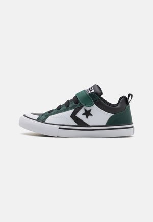 Zapatilla con parte superior de piel blanca, detalles en negro y verde, puntera redondeada, cordones negros y una correa de velcro en el tobillo. Suela de goma.