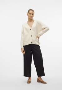 Creme cardigan med V-hals, knaplukning foran og to forlommer. Matched med sorte brede bukser og brune flade sko. Glat tekstur.