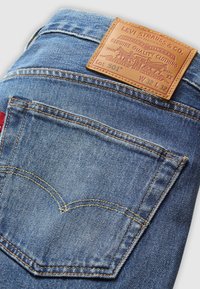 Jean Levi's 501 en denim bleu plié, montrant la poche arrière, les surpiqûres orange, l'étiquette rouge et l'étiquette en cuir avec la taille W32 L32.