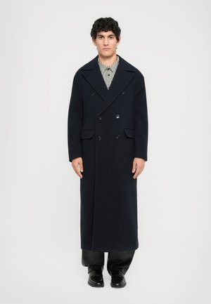 Man staat, gekleed in een lange zwarte dubbelzijdige jas over een geruit shirt, zwarte broek en zwarte schoenen, tegen een effen witte achtergrond.