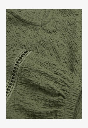 Close-up af olivengrønt tekstureret stof med rynket mønster og en dekorativ blonderkant detalje på ærmet.