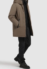 Chaqueta acolchada marrón con capucha, con cierre de cremallera y botones, dos bolsillos laterales y un diseño elegante. Llevada sobre una capa negra.