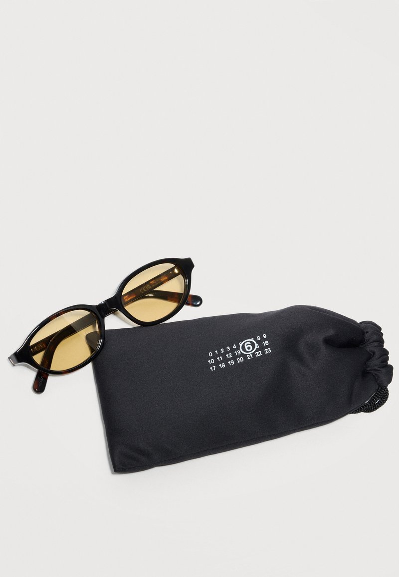 Lunettes de soleil à monture ovale noire avec verres ambrés reposant à côté d'une pochette souple noire comportant des numéros imprimés en blanc et un 6 encerclé.