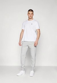 Witte T-shirt met korte mouwen en een klein logo, gecombineerd met lichtgrijze joggingbroek en witte sneakers, staand tegen een neutrale achtergrond.