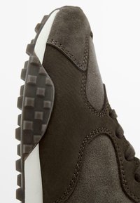 Tmavě hnědé semišové tenisky s robustní gumovou podrážkou, polstrovaným límcem a kontrastními bílými prvky podél boku.