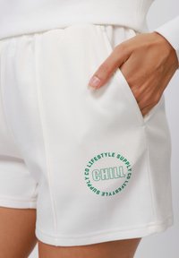 Valkoiset shortsit, jotka on valmistettu pehmeästä kankaasta. Niissä on vihreä logo, jossa lukee "CHILL" lahkeessa, sekä sivutaskut joustavalla vyötäröllä.