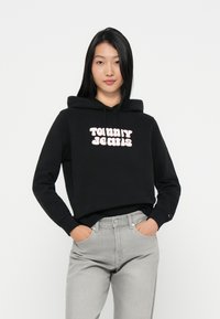 Sweatshirt cropped preto com o logotipo rosa "Tommy Jeans" na parte da frente. Combina com jeans cinza claro, exibindo um ajuste casual e descontraído.