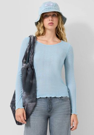 Jonge vrouw draagt een lichtblauw langarmshirt, bijpassende bucket hat, grijze jeans, met aan haar schouder een grote donkere tie-dye tas.