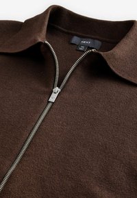 Pull en tricot brun à fermeture éclair avec col, doté d'une fermeture éclair en métal argenté et d'un tissu texturé. Étiquette indiquant "NEXT", taille moyenne.