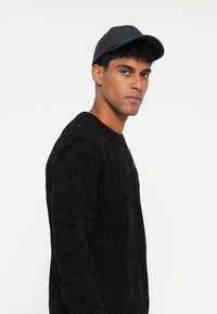 Schwarzer gestrickter Pullover mit strukturierten Mustern, kombiniert mit einer schwarzen Baseballkappe. Das Design verfügt über lange Ärmel und einen runden Ausschnitt.