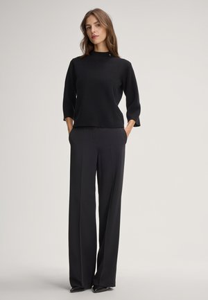 Pull noir à texture avec un col haut, associé à un pantalon large noir. Les deux vêtements sont sans couture et au design minimaliste.