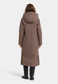 Brauner gepolsterter langer Mantel mit Kapuze, der eine taillierte Taille und seitliche Schlitze aufweist, über schwarzen Hosen und Stiefeln getragen, von hinten gezeigt.